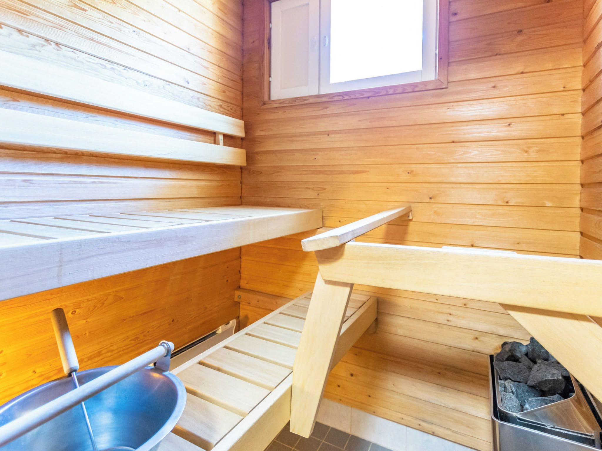 A sauna room with wooden benches, a window, and a metal container.