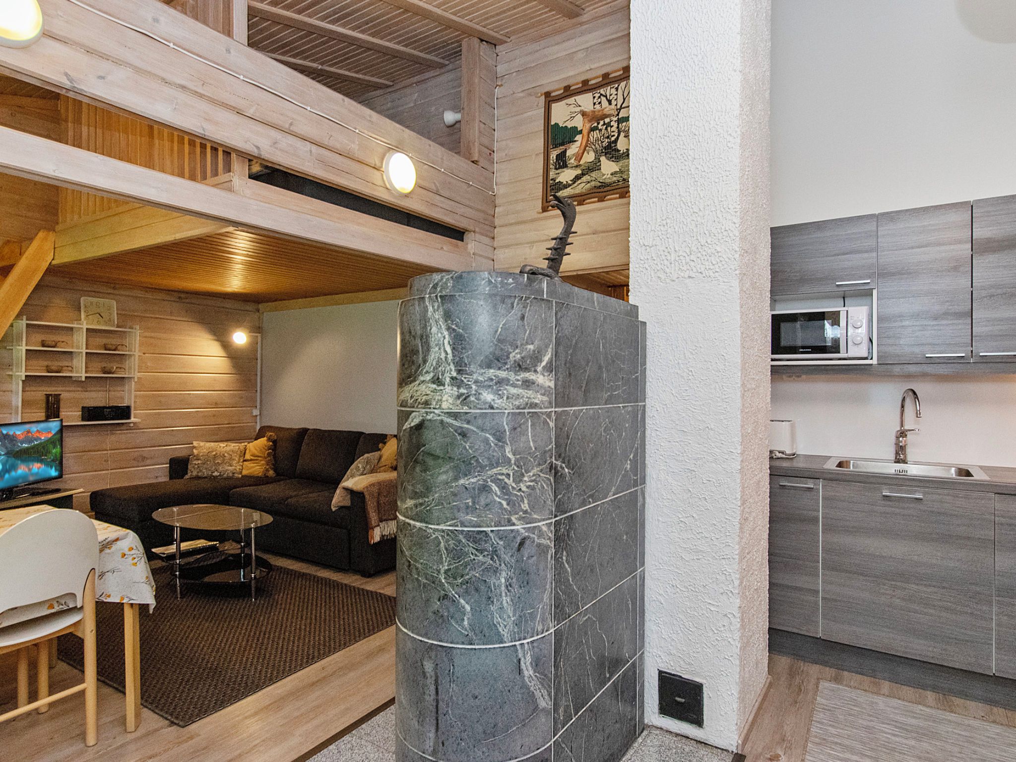 Interior moderno com um pilar de pedra, vigas de madeira no teto, um sofá, uma televisão e uma área de cozinha.