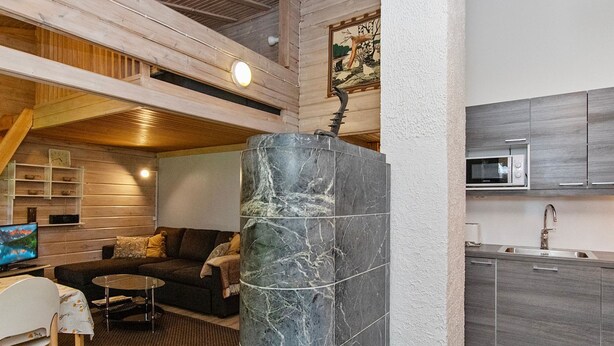 Een modern interieur met een stenen zuil, houten balken in het plafond, een keuken met grijze kastjes, en een zithoek met een bank en televisie.