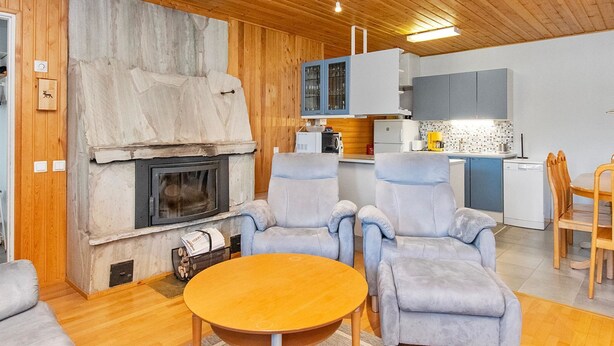 Ein modernes Wohnzimmer mit Kamin aus Stein, grauen Sofas, einem Holztisch und einer Küchenzeile mit blauen Schränken und einem weißen Kühlschrank.