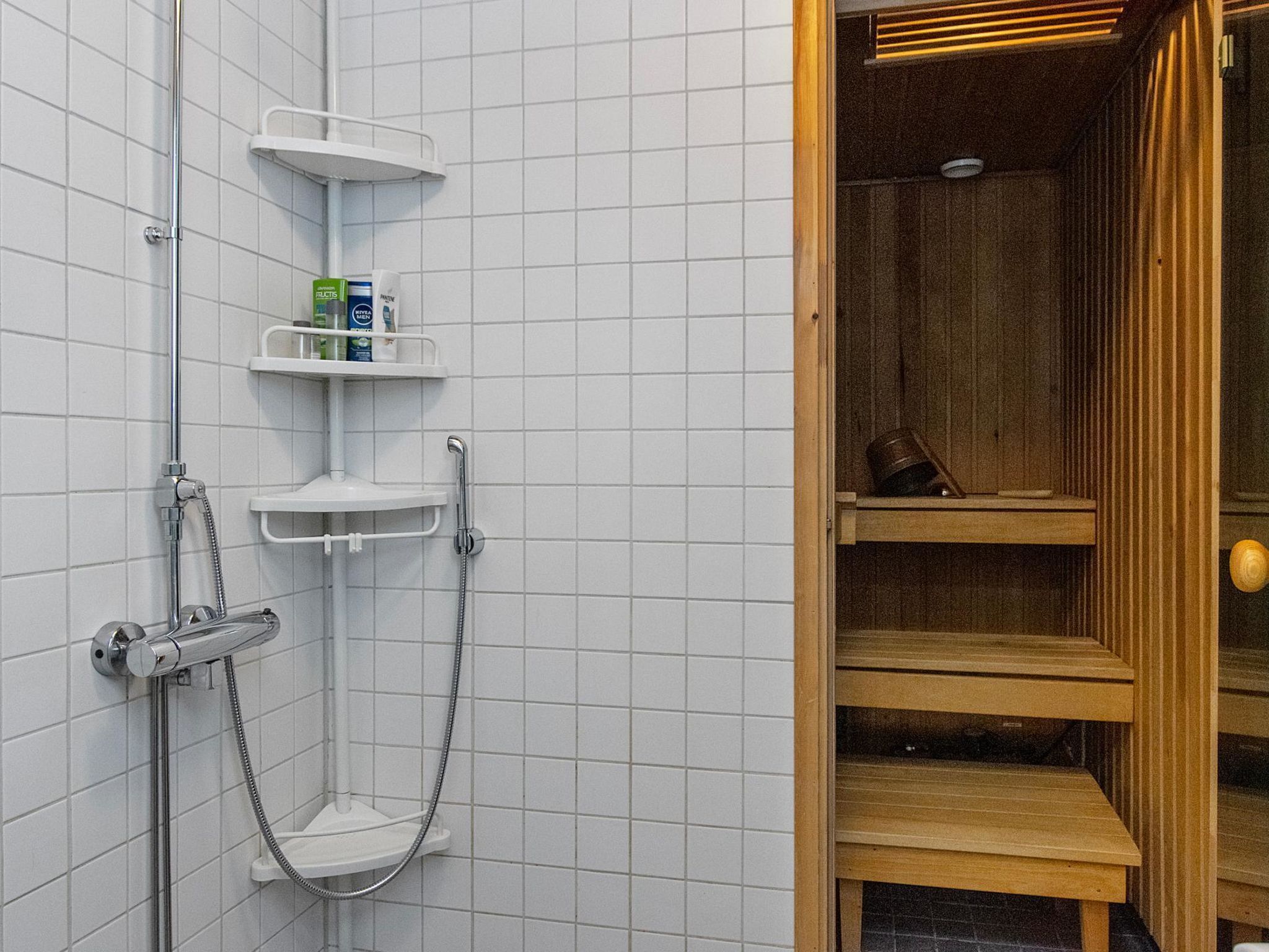 Suihkualue, jossa on krominen suihkupää, käsisuihkupää ja lasiseinällä varustettu suihkukaappi. Seinät ovat valkoisilla laatoilla päällystettyjä, ja ovikehykset ovat puuta.