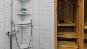 Ein Duschbereich mit Duschkopf aus Chrom, einer Handbrause und einer gläsernen Duschkabine. Die Wände sind mit weißen Fliesen versehen und der Türrahmen ist aus Holz.