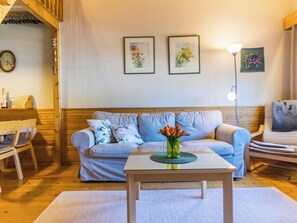 Un salon chaleureux avec un plancher en bois, un canapé bleu clair agrémenté de coussins à motifs, une petite table basse en bois sur laquelle est posé un vase de fleurs, une table d’appoint avec une lampe, et des œuvres d’art encadrées accrochées au mur.