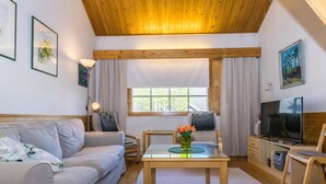 Un salón acogedor con techo de madera, un sofá, una mesa de centro con flores, un televisor y una ventana con cortinas.