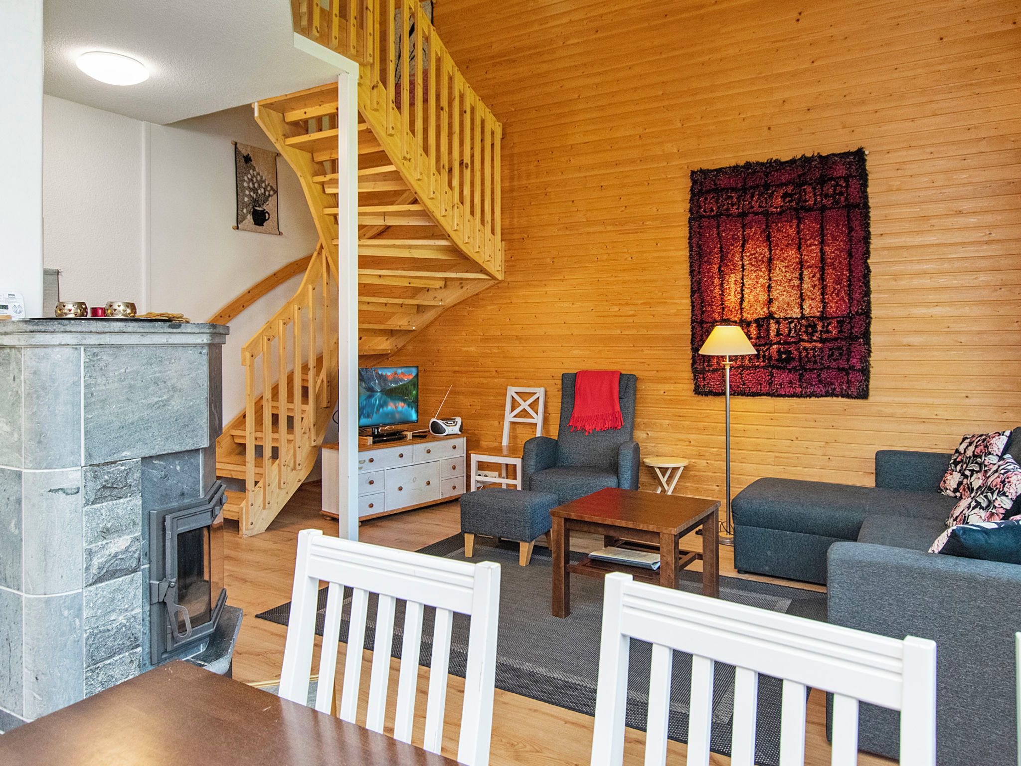 A cozy living room with a fireplace, wooden walls, a staircase, and a large tapestry.