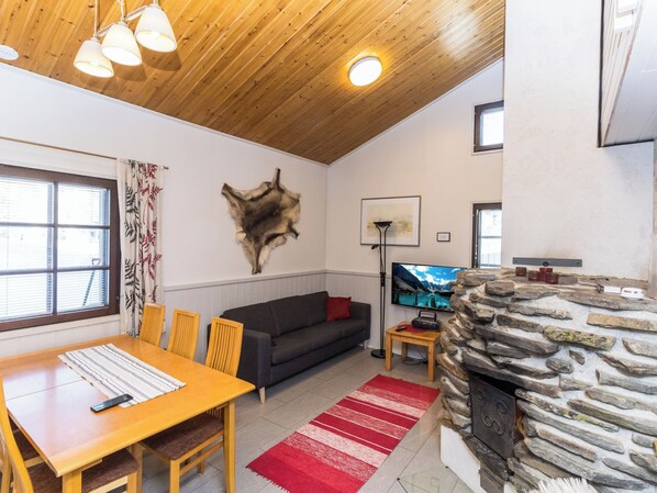 A cozy living room with a stone fireplace, wooden ceiling, and a large window with curtains.