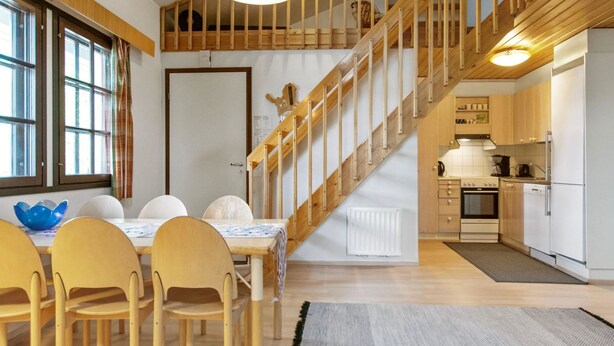 Cocina moderna con detalles de madera, zona de comedor con mesa y sillas, y una escalera que sube a un nivel superior.