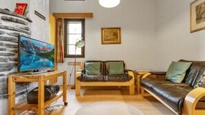 Una sala de estar acogedora con un sofá de madera, un televisor sobre un mueble de madera y un ventanal con cortinas.