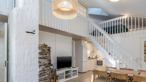 A spacious living area with a high ceiling, a stone wall, a fireplace, a TV, a dining table, and a staircase leading to an upper level. - Saarihely a1 by Interhome (Inari)