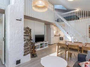 Amplia sala de estar con techo alto, pared de piedra, chimenea, televisor, mesa de comedor y escalera que conduce a un nivel superior.