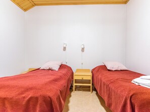 A simple room with two beds, each with a red blanket and a pillow, a small wooden nightstand, and a white wall.