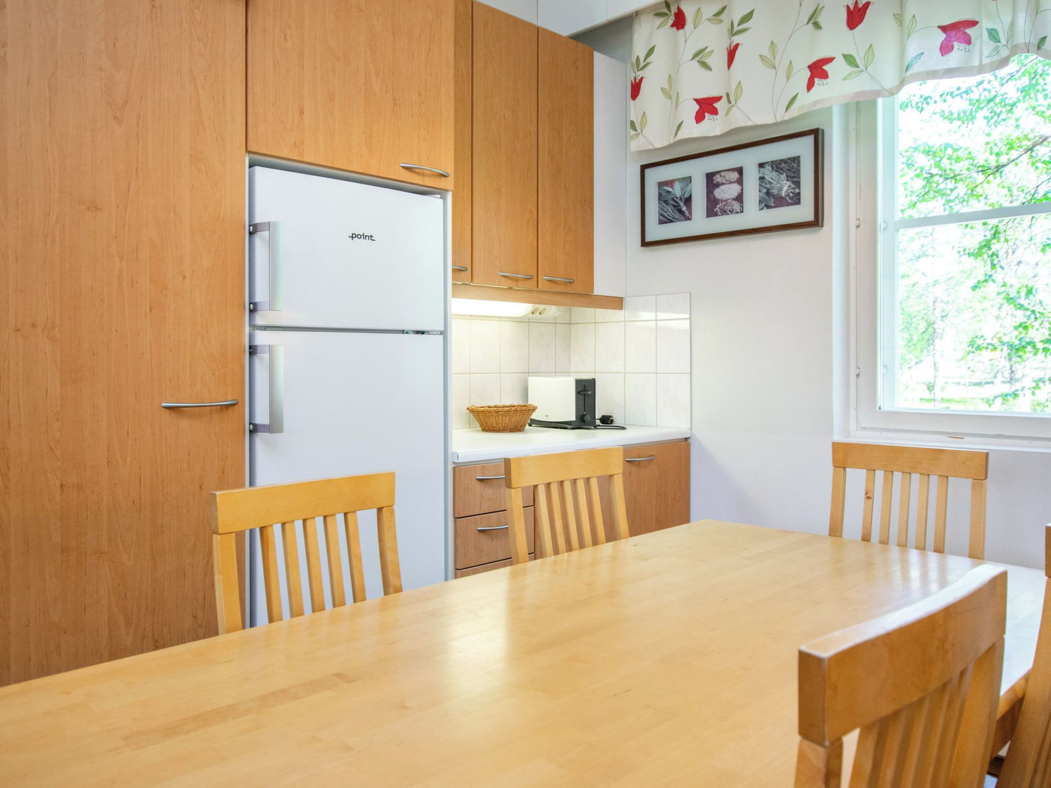 Une cuisine avec des meubles en bois, un réfrigérateur blanc, une table à manger avec des chaises et une fenêtre donnant sur des arbres.