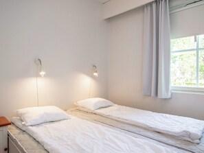 A simple, clean room with two single beds, a window with curtains, and wall-mounted lights.