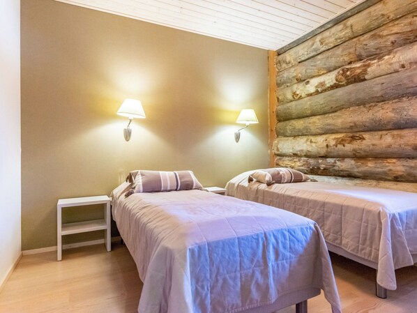 A room with two beds, each with a pillow and a bedside table. There are wall-mounted lamps on either side of the beds. The room has a wooden log wall on one side and a plain wall on the other.
