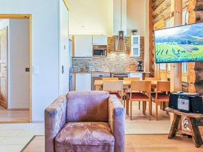 A modern living room with a leather armchair, a dining area with wooden chairs, a kitchen with stone backsplash, and a flat-screen TV.