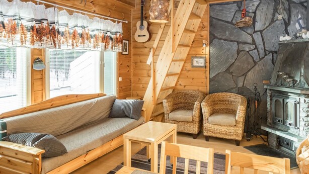 L’intérieur chaleureux d’un chalet avec un canapé en bois, des fauteuils en osier, une cheminée en pierre et une guitare accrochée au mur.