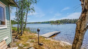 Una cabana al costat d'un llac amb un moll que s'endinsa a l'aigua, envoltada d'arbres i un cel serè.