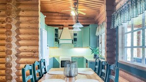 Interior d'una cabana de fusta amb taula de dinar, cadires, zona de cuina i finestra amb cortines.