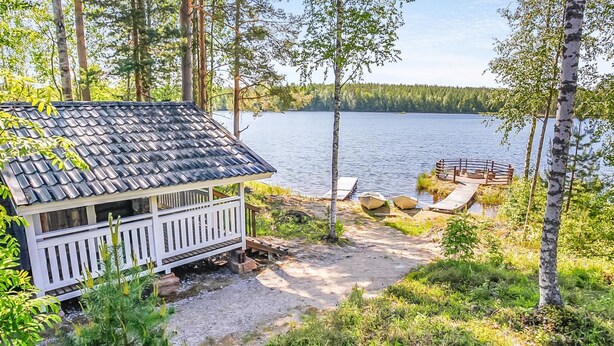Une cabane avec un toit en tuiles, une terrasse et un quai s’avançant dans un lac entouré d’arbres.