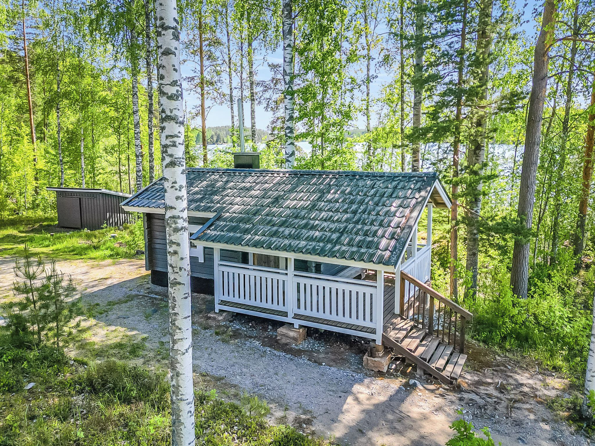 A cabin with a green roof, white railings, and a gravel driveway surrounded by trees.