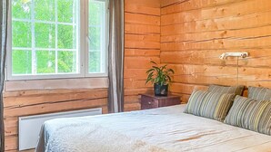 Une chambre dans une cabane en bois, avec une grande fenêtre, un radiateur, une table de chevet agrémentée d’une plante et des oreillers à rayures.