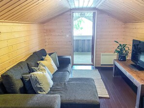 A cozy cabin interior with a wooden ceiling, a sofa with cushions, a television on a wooden table, and a sliding door leading to an outdoor area.
