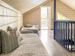 A loft bedroom with a high ceiling, a wooden sloped roof, a bed with a quilted mattress, a bench with pillows, and a black metal railing.