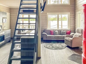 Una escalera conduce a una zona de altillo, una sala de estar con sofá y sillones, y una cocina con armarios y un refrigerador.