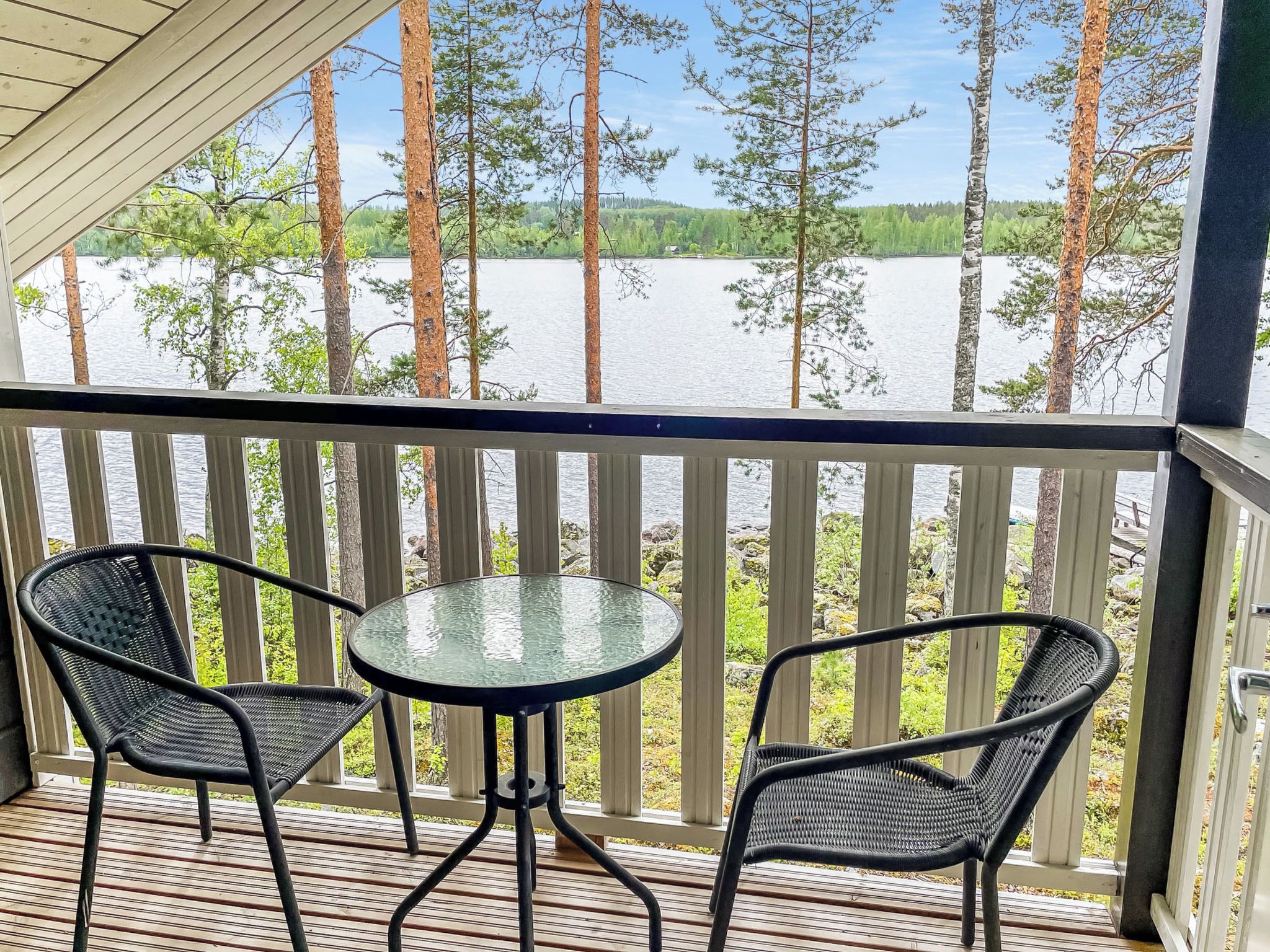 Ett takförsett utomhusområde med ett bord och två stolar, med utsikt över en vattenyta och ett skogsområde.
