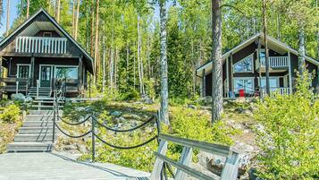 A wooden cabin with a deck leading to the entrance, surrounded by a forest.