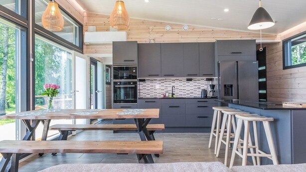 Una cocina moderna con armarios oscuros, electrodomésticos de acero inoxidable y una isla de madera. Cuenta con ventanas amplias en un lateral y una zona de comedor con mesas y bancos de madera.