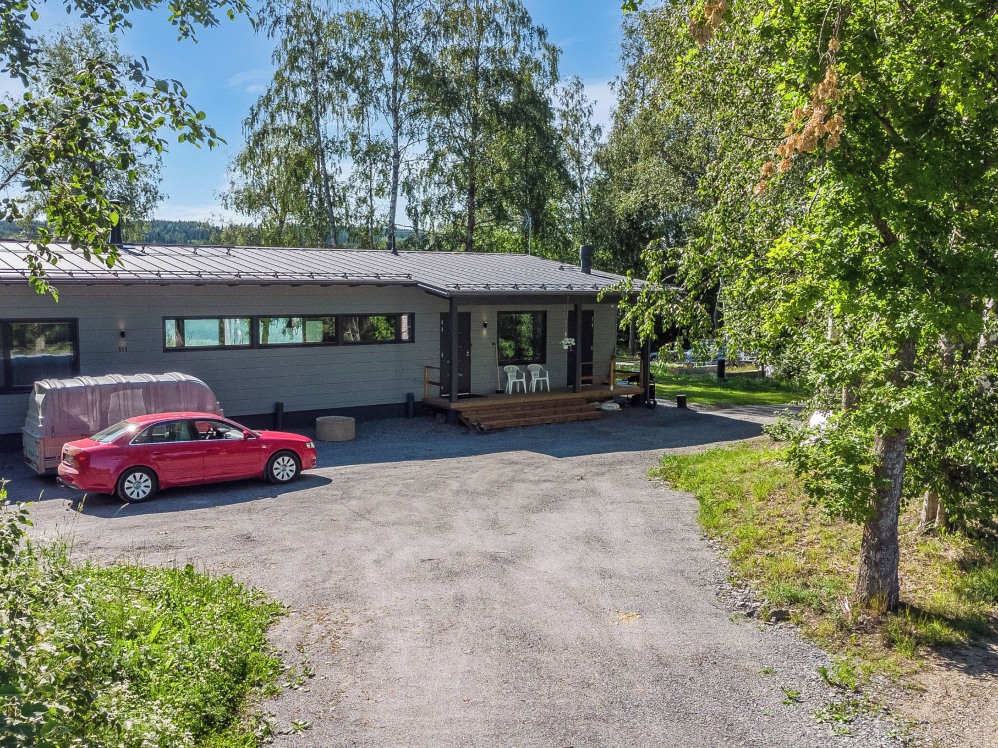 Yhden kerroksen talo, jossa on katettu kuisti, sorapäällysteinen ajotie ja punainen auto pysäköitynä eteen.