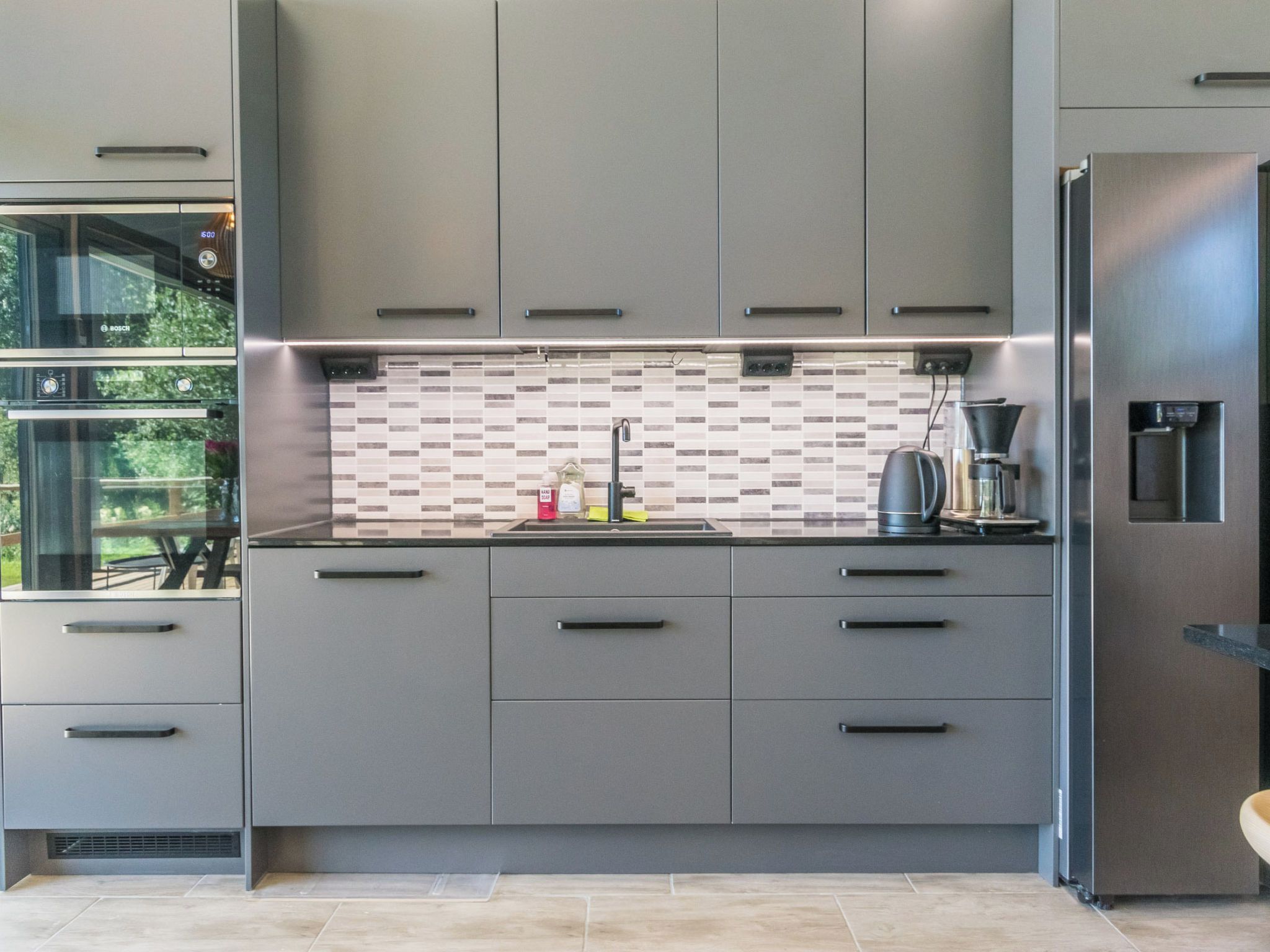 A modern kitchen with grey cabinets, a stainless steel refrigerator, and a tiled backsplash.