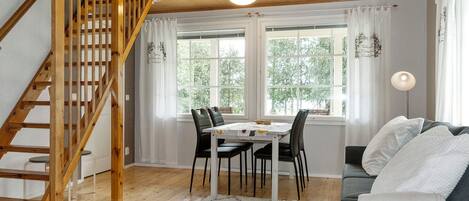 Un salon chaleureux avec un escalier en bois, un coin repas avec une table et des chaises, et un canapé agrémenté d’un plaid.