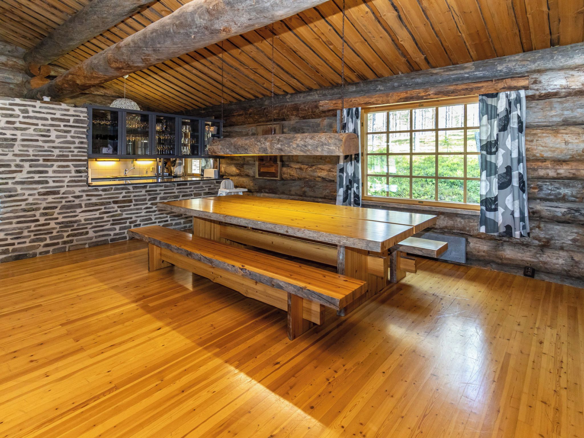 Un intérieur rustique en bois, avec une grande table à manger, des murs en pierre et une vue sur la verdure environnante.