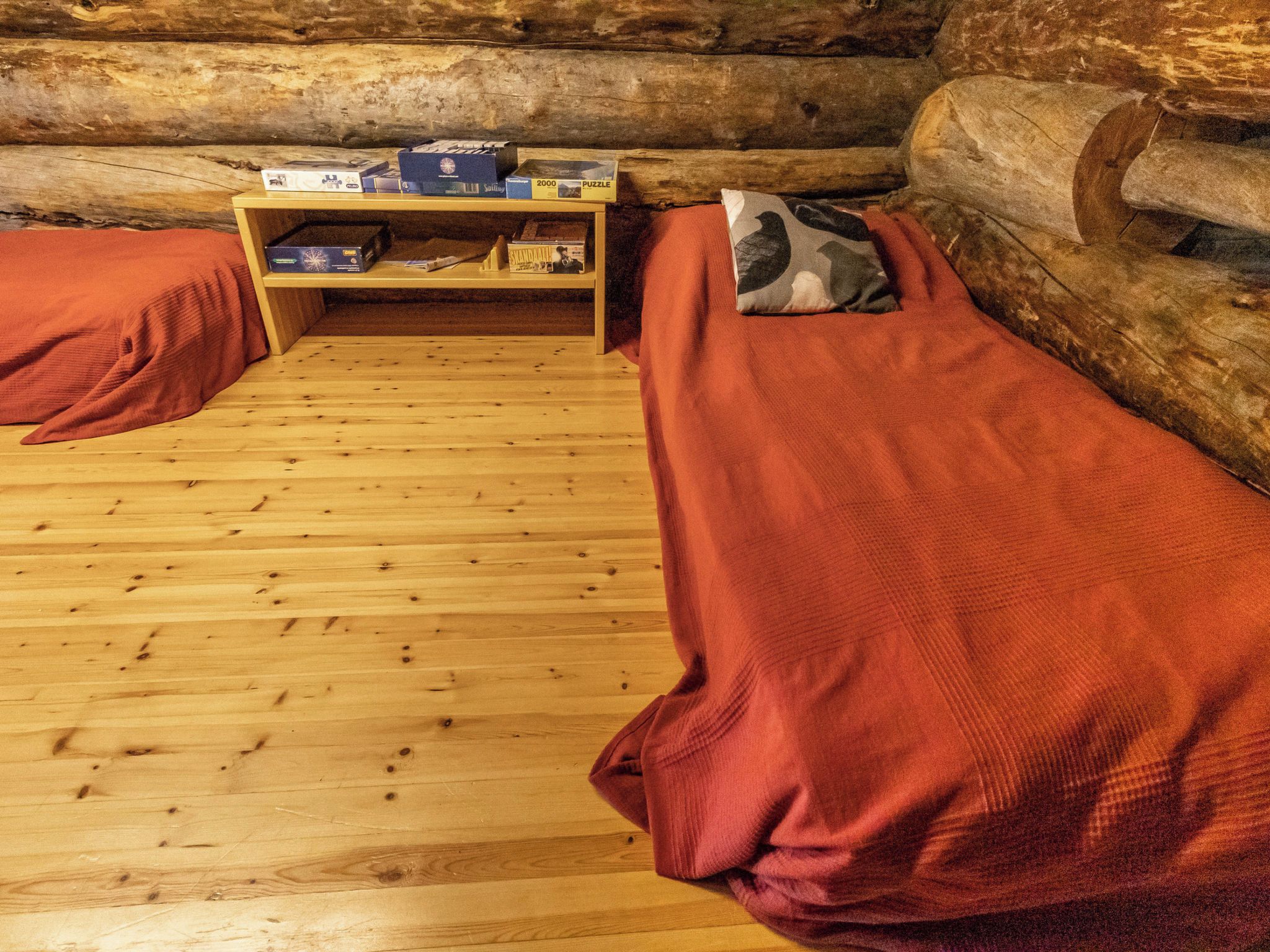 A log cabin room with two beds, a wooden floor, and a shelf with books and puzzles.