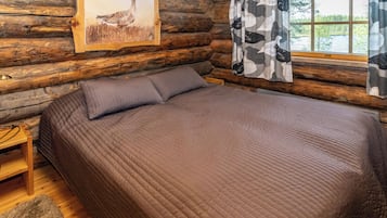 Ein Schlafzimmer in einer Blockhütte mit einem Bett, Kissen, einem Gemälde und einem Fenster mit Vorhängen.