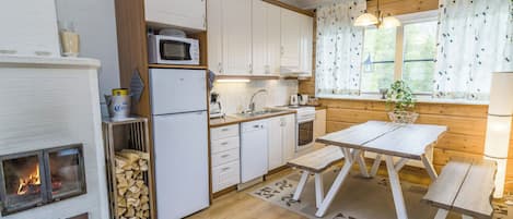 Une cuisine compacte équipée d’un four à micro-ondes, d’un réfrigérateur et de meubles en bois. On y trouve une table à manger avec des bancs, ainsi qu’une cheminée avec un feu de bois.