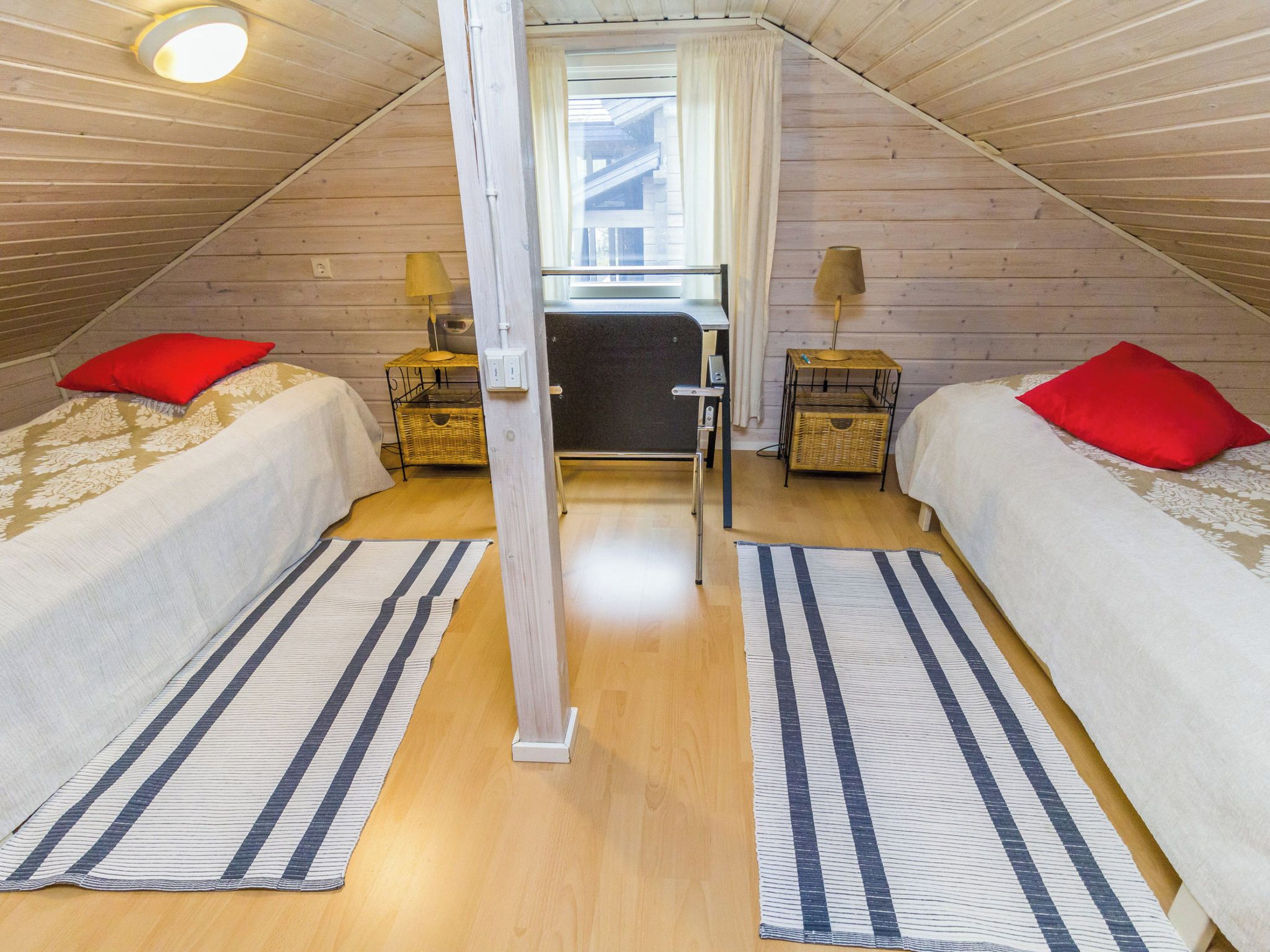 Une chambre avec deux lits, chacun agrémenté d’un oreiller rouge, et une fenêtre avec des rideaux. On y trouve deux lampes sur des tables de chevet, ainsi qu’un plancher en bois.