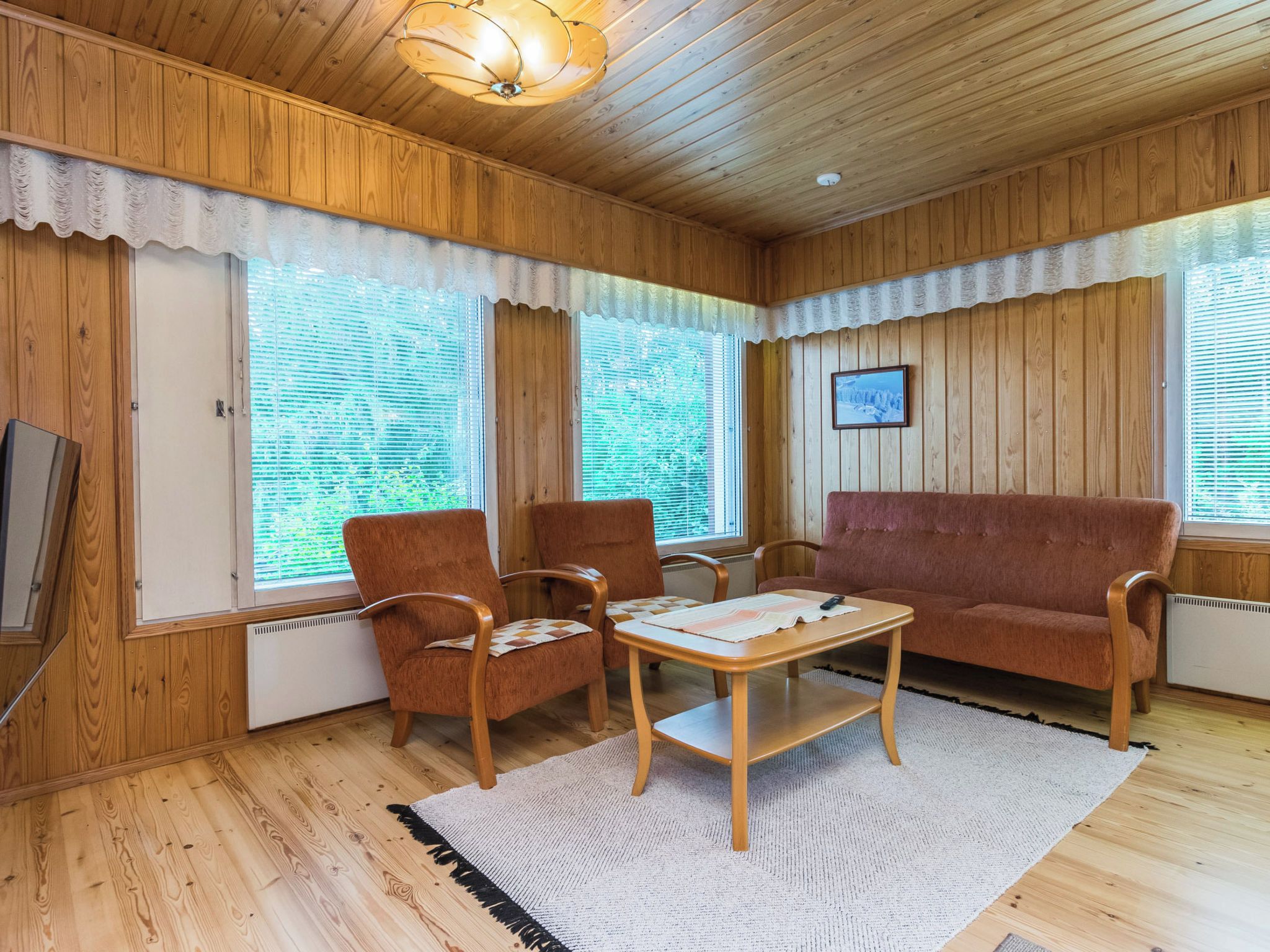 Ein gemütliches Zimmer mit Holzwänden und -decke, einer Couch, zwei Sesseln, einem kleinen Tisch und einem großen Fenster mit Jalousien.