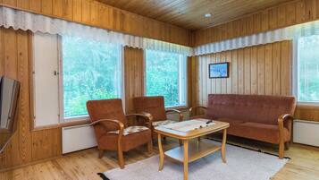 Ein gemütliches Zimmer mit Holzwänden und -decke, einer Couch, zwei Sesseln, einem kleinen Tisch und einem großen Fenster mit Jalousien.