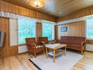 Una habitación acogedora con paredes y techo de madera, un sofá, dos sillones, una mesita y un ventanal grande con persianas.
