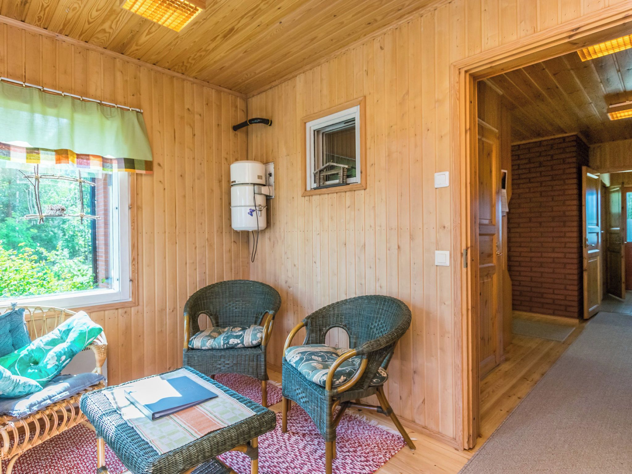 A wooden room with two wicker chairs, a small table with a book, a window with a curtain, and a heater on the wall.