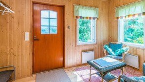 Une pièce chaleureuse avec des murs en bois, une porte en bois, deux fenêtres avec des rideaux, un fauteuil en osier, une petite table et un radiateur.