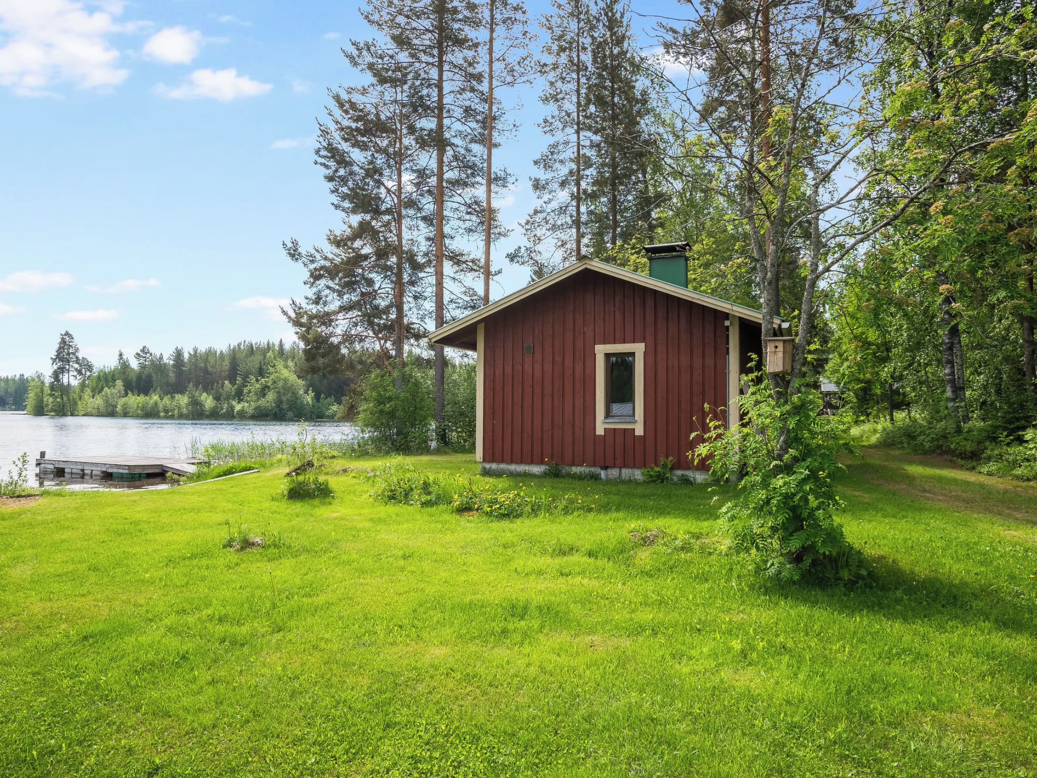 A small red cabin with a dock extending into a lake, surrounded by dense forest.
