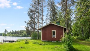 Une petite cabane rouge, dotée d’une fenêtre et d’une porte, entourée d’une zone herbeuse et de grands arbres, près d’une étendue d’eau.