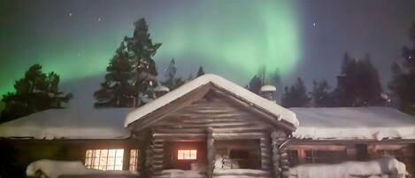En træhytte med snebetaget tag og et oplyst vindue, omgivet af sne og træer under en nattehimmel med nordlys.