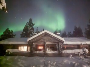 En træhytte med snebetaget tag og et oplyst vindue, omgivet af sne og træer under en nattehimmel med nordlys.