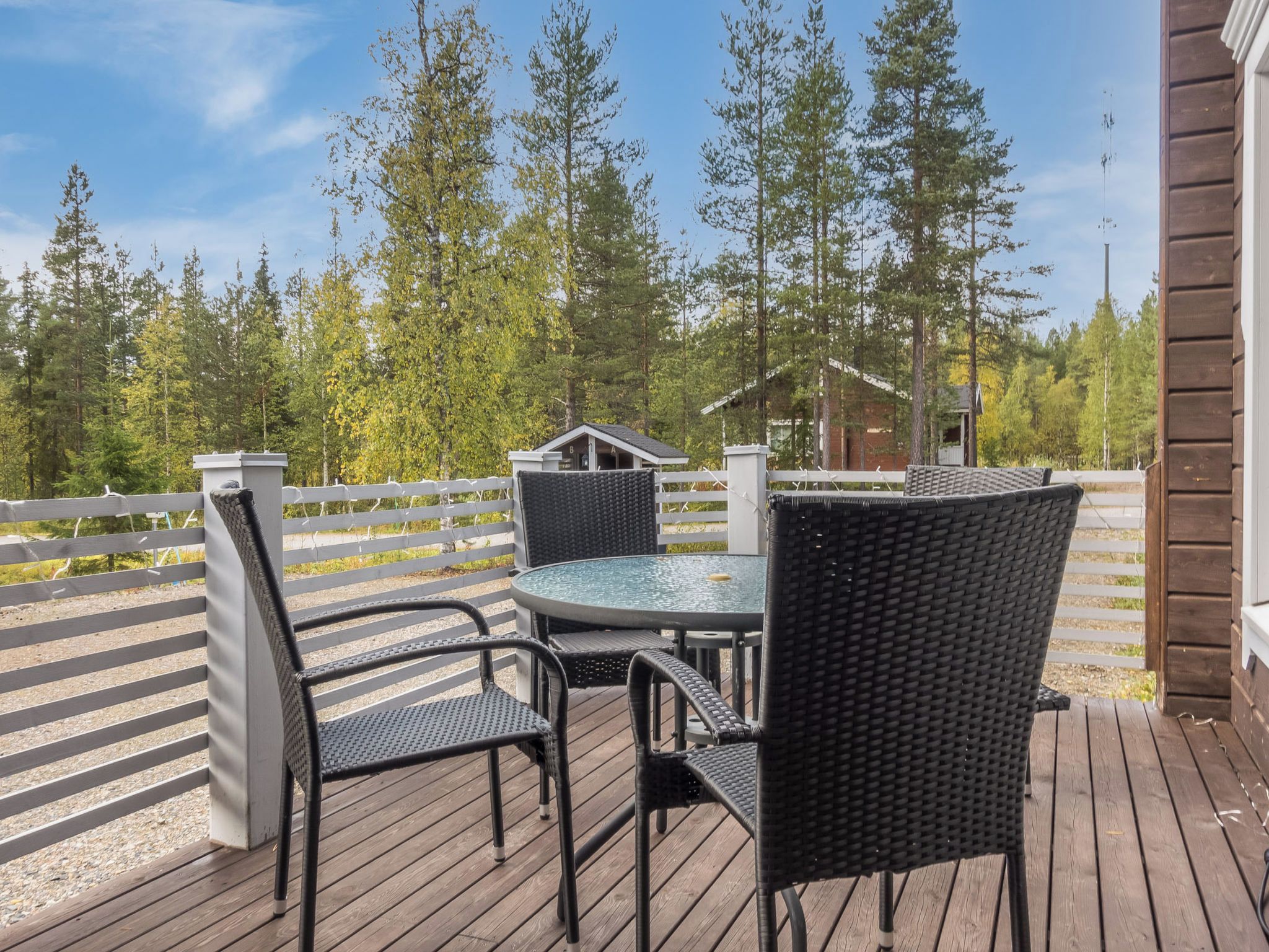 Une terrasse en bois avec une table ronde et quatre chaises, entourée d’une clôture blanche et de grands arbres.
