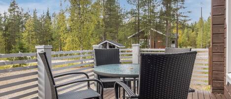 Eine Holzterrasse mit einem runden Tisch und vier Stühlen, umgeben von einem weißen Zaun und hohen Bäumen.